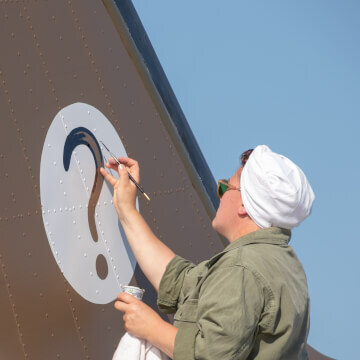 A women paints the 319th Troop Carrier Squadron insignia (a question mark) on the vertical stabilizer of ‘Hairless Joe’, a Douglas C-47 Skytrain.